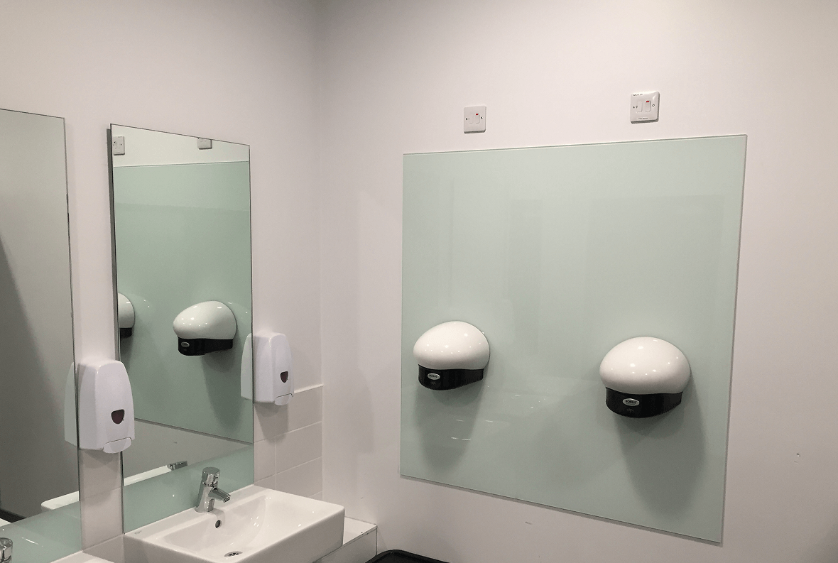 White Splashback In Wc Restroom With White Hand Dryers Mounted On A Light Green Glass Panel.